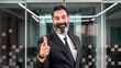 © HI Pictures - A middle-aged, dark-haired, bearded businessman is standing in an empty office environment with a blurred background, smiling at the camera while pointing towards it with his index finger