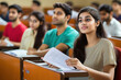© Neha - Indian college students in a lecture hall, sitting at desks and listening to the teacher while holding papers or notes