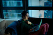 © Connect Images - Woman in glasses using a laptop while sitting on the floor by a window at dusk.