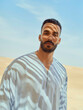 © Connect Images - A serene man in traditional attire stands against a desert backdrop, with a clear blue sky overhead, Morocco