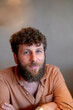 © Connect Images - A smiling bearded man with curly hair wearing a salmon-colored shirt posing for a portrait with a blurred background.