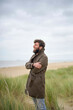 © Connect Images - Smiling bearded man standing with arms crossed in a coastal grassland, wearing a trench coat, Belgium