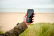 © Connect Images - Hand holding a smartphone with a compass app on the screen at a sandy beach under a cloudy sky, Belgium