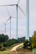 © yaqui_villegas - A close-up perspective showcasing several tall wind turbines, positioned along a dirt path, surrounded by greenery and trees, under a cloudy sky in a wide open space in Navarra Spain