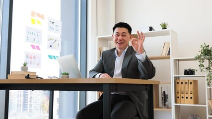 Wall Mural - Portrait of Asian male businessman in 30s working with laptop in modern office. He is smiling and making an okay gesture, showcasing confidence and positive attitude.