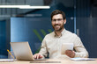 © Tetiana - Portrait of a smiling young man in glasses sitting in the office at a desk with a notebook, holding a notebook and looking at the camera