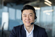 © Tetiana - Close-up portrait of a young Asian man in a suit and headset in the office, sitting in front of the camera and smiling