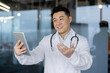 © Tetiana - Smiling young Asian male doctor standing inside hospital, holding tablet, talking and consulting on video call