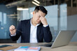 © Tetiana - Tired young Russian man sits in the office at the workplace in front of a laptop, holds glasses and rubs his eyes with his hand