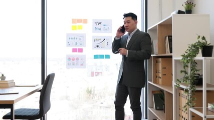 Wall Mural - Asian businessman in suit stands in modern office engaged with graphs and charts on window. He communicates with colleague via smartphone highlighting multitasking and professionalism in workspace.