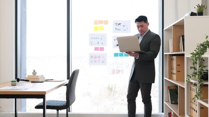 Wall Mural - Asian businessman in formal suit discusses graphs and charts during online video call. Standing by panoramic window in modern office, he uses laptop for engaging presentation.