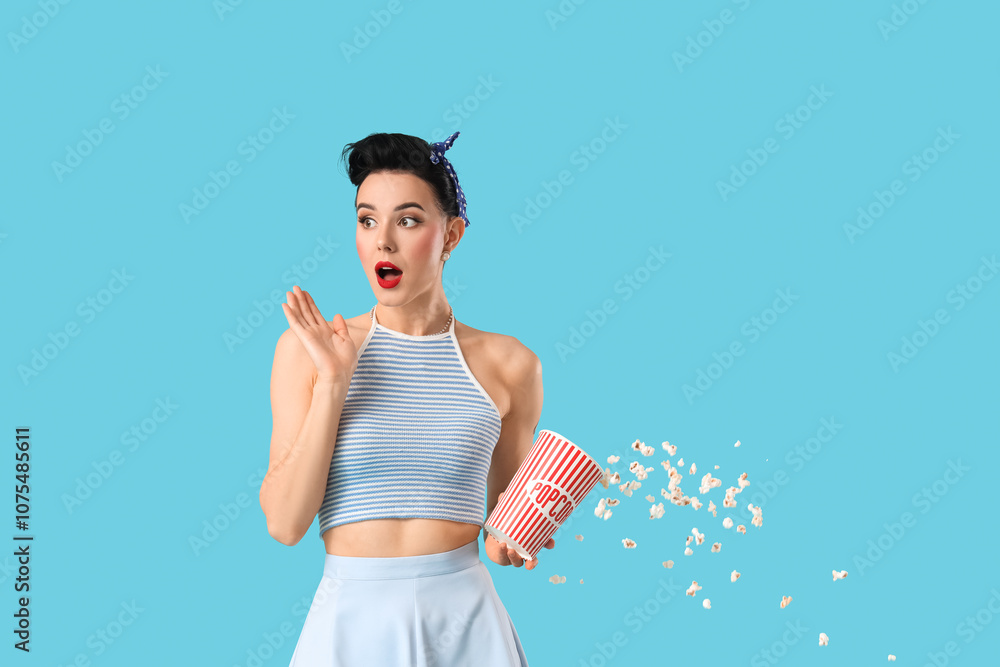 Shocked young pin-up woman spilling popcorn on blue background