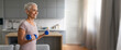 © Prostock-studio - A smiling older woman focuses on her workout, lifting blue dumbbells in a well-lit kitchen. The cozy environment reflects her commitment to fitness and health.