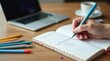© Alex Pios - A close-up view of a hand writing notes in a notebook with colorful pens scattered around.
