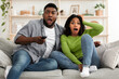 © Prostock-studio - Coronavirus News. Shocked african american couple watching tv with open mouth, staying at home together, sitting on sofa in living room, free space