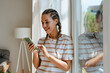 © Westend61 - Smiling young woman using smart phone near window at home