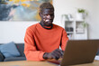 © Studio Marmellata - A young man with vitiligo dressed in an orange shirt types on a laptop while seated at a desk in a bright office with modern furniture and artwork, surrounded by a relaxed and professional atmosphere.