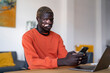 © Studio Marmellata - man with vitiligo dressed in an orange shirt sits at a desk while looking intently at his smartphone, surrounded by greenery and furniture, in a modern indoor environment with natural light
