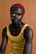 © Studio Marmellata - Black man with vitiligo wearing a yellow tank top and a red beanie sits against a brown background, looking into the camera with a calm and contemplative expression, showcasing individuality and style