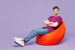 © ViDi Studio - Full body young smiling happy man wear violet t-shirt casual clothes sit in bag chair hold hands crossed folded look camera isolated on plain pastel light purple background studio. Lifestyle concept.