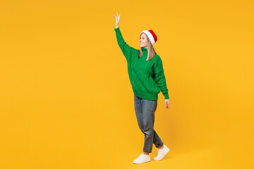 Wall Mural - Full body side view merry smiling young woman wear green casual sweater Santa hat posing walk go waving hand isolated on plain yellow background. Happy New Year, celebration Christmas holiday concept.