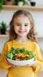 © Sergio - A cheerful girl in a yellow shirt holding a fresh salad in a cozy kitchen with plants in the background during daylight