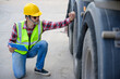 © เลิศลักษณ์ ทิพชัย - A worker wearing a safety helmet, reflective vest, and holding a clipboard performs a tire inspection on a large truck. which is perfect for logistics and industrial projects.