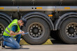© เลิศลักษณ์ ทิพชัย - A safety inspector inspects the tires of a big fuel tanker while wearing a reflective vest. He maintains adherence to transportation safety regulations while holding a clipboard.