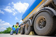 © เลิศลักษณ์ ทิพชัย - A safety inspector in a reflective vest checks the tires of a large fuel tanker under a bright blue sky. Holding a clipboard, he ensures compliance with transportation safety standards.