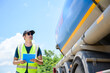 © เลิศลักษณ์ ทิพชัย - A logistics worker in a reflective vest holds a clipboard as he inspects a large fuel tanker on a sunny day. The image highlights themes of transportation and safety compliance.