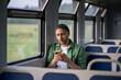 © DimaBerlin - Relaxed man wearing wireless earphones, using smartphone and surfing Internet, social media in train car. Tech enhanced journey, tourist commuting experience, public transit with music and podcasts