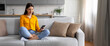 © Prostock-studio - A young woman sits cross-legged on a comfortable sofa, smiling as she interacts with her smartphone.
