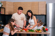 © dvulikaia - Young parents teach their little daughters to cook vegetarian salad at the kitchen.