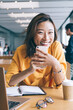 © BullRun - Portrait of cheerful Asian female blogger with cellphone gadget in hands smiling at camera during e learning time, successful Chinese woman rejoicing near desktop with modern laptop computer