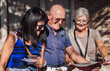 © luciano - Joyful group of senior friends visiting Alhambra in Granada listening to information via audio guide. Elderly man and two women enjoying vacation and retirement traveling in Europe