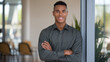 © Tom - Smiling young Black man in a grey shirt, standing confidently with arms crossed in a modern office setting.