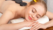 © Prostock-studio - Health and beauty concept. Smiling woman with closed eyes with hot stones on back lie on white towel and on massage table