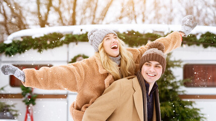 Wall Mural - A cheerful couple plays in the snow, with the woman joyfully riding on the man's back. Lush trees and a cozy holiday setting create a festive atmosphere.