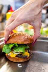 Wall Mural - Close-up of hand with purple nail polish placing the top bun on cheeseburger, highlighting fresh ingredients and fast food preparation. Perfect for food industry, culinary arts, and fast food themes.