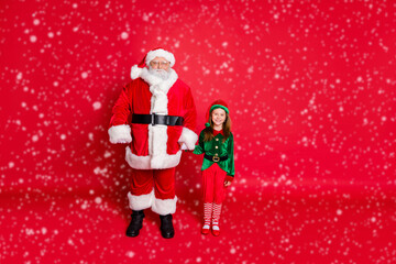 Wall Mural - Full size photo of cheerful two santa and elf holding hands ready to enjoy newyear noel wearing bright green costume isolated over red background