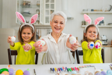 Wall Mural - Happy Easter. Two little girls sisters twins grandmother painting eggs enjoying time together. Happy family grandma granddaughters child kids preparing for Easter. Spring Christian festival tradition