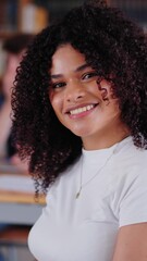 Wall Mural - Vertical. High school girl poses looking smiling at camera sitting in library with classmates in background. Group friends meeting together in study room. Portrait happy young Latin female student