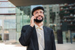 © Jose Calsina - enthusiastic young bearded entrepreneur enjoying meaningful business conversation on his smartphone while standing outside corporate building, showcasing his confident and approachable demeanor during