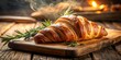 © Indigo Studio - A freshly baked croissant with a warm, golden hue, resting on a rustic wooden board, adorned with sprigs of rosemary, emitting a tantalizing aroma of buttery delight.