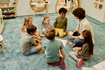 group of cute intercultural kids sitting in circle on the floor and looking at young female teacher 
