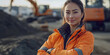© VALUEINVESTOR - A young and beautiful female worker, wearing an orange jacket with her arms crossed in front of a construction site background, generative AI