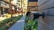 © Thanakrit - A security camera is mounted on a wooden wall, overseeing a well-maintained pathway lined with plants and modern residential buildings in the background.