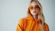 © Helois - Stylish blonde woman wearing orange hoodie and sunglasses against a white background.