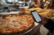 © dyshlivenko - A person is holding a phone while admiring a large pizza with melted cheese at a lively pizzeria. The kitchen is busy with chefs preparing various dishes in the background