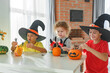© Olga Gimaeva - Three real siblings two brothers and sister making together jack o lantern at their home white kitchen, cutting real pumpkins and playing with paper black ghosts, lovely vibe of halloween